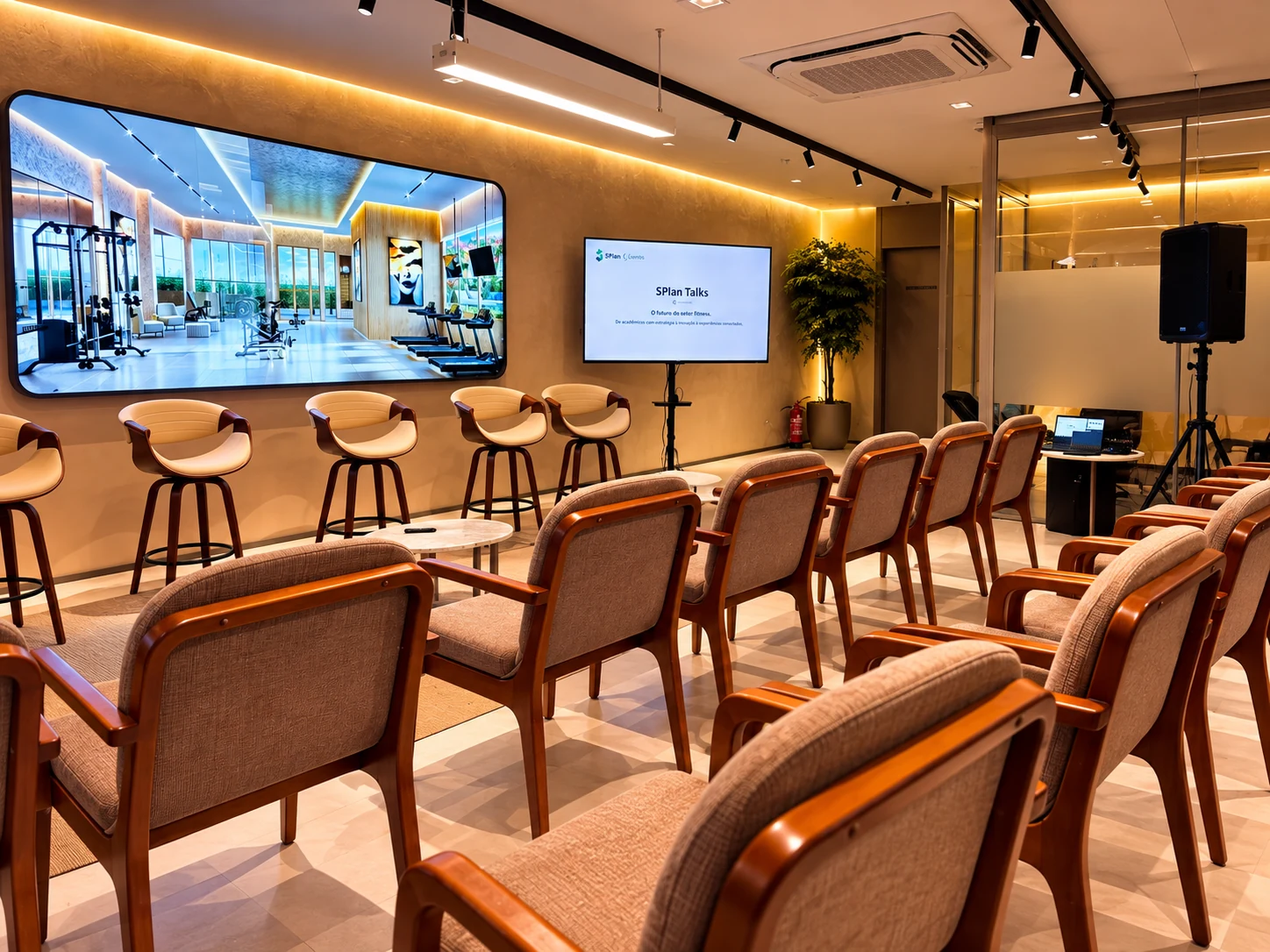 TV em pedestal exibindo apresentação em evento corporativo, com plateia, caixa de som e notebook de operação